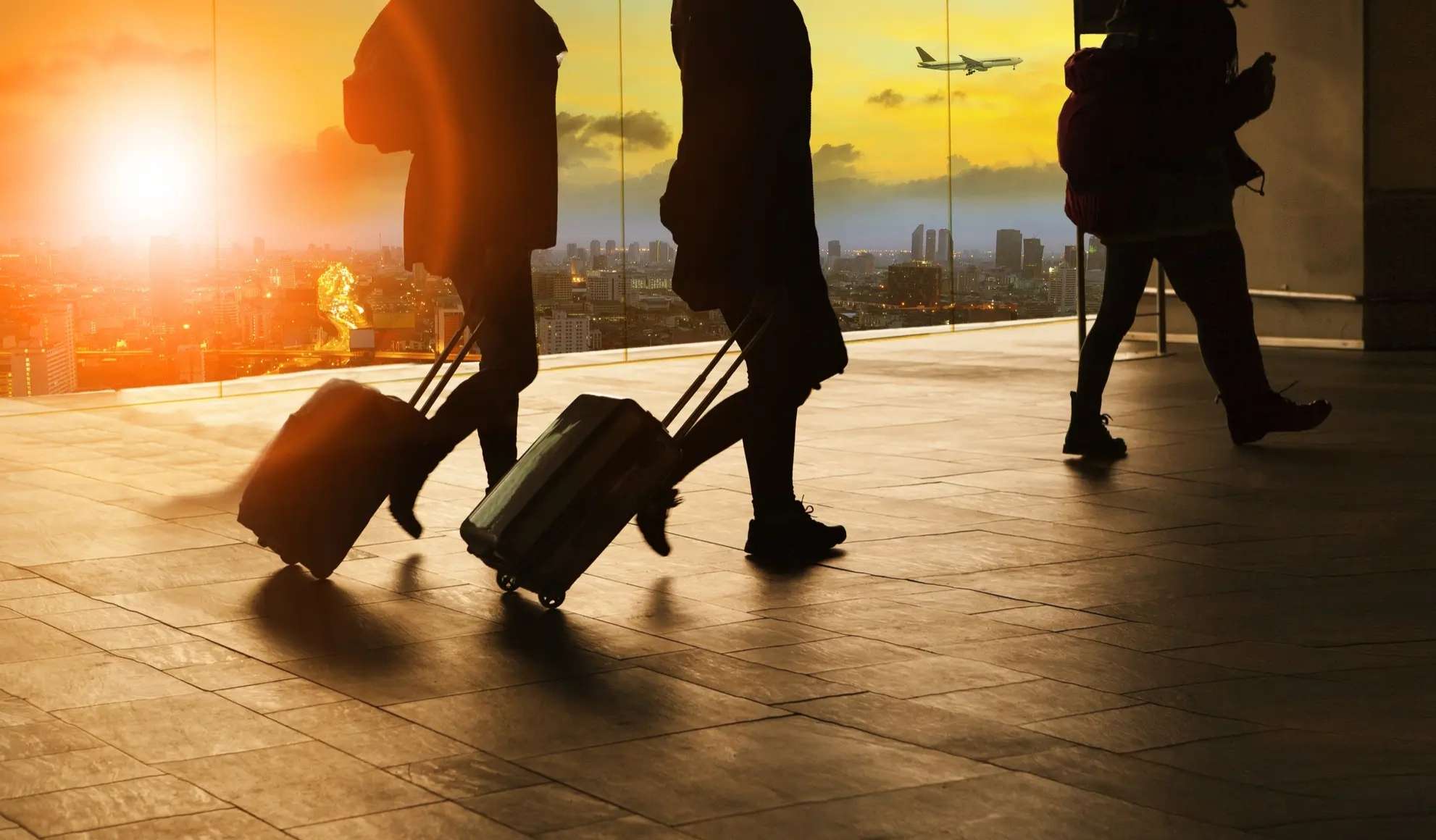 quanto-costa-noleggiare-un-aereo-charter-flyness Passeggeri in aeroporto con trolley con aereo in decollo sullo sfondo, simbolo di organizzazione di un volo charter. Flyness air charter broker