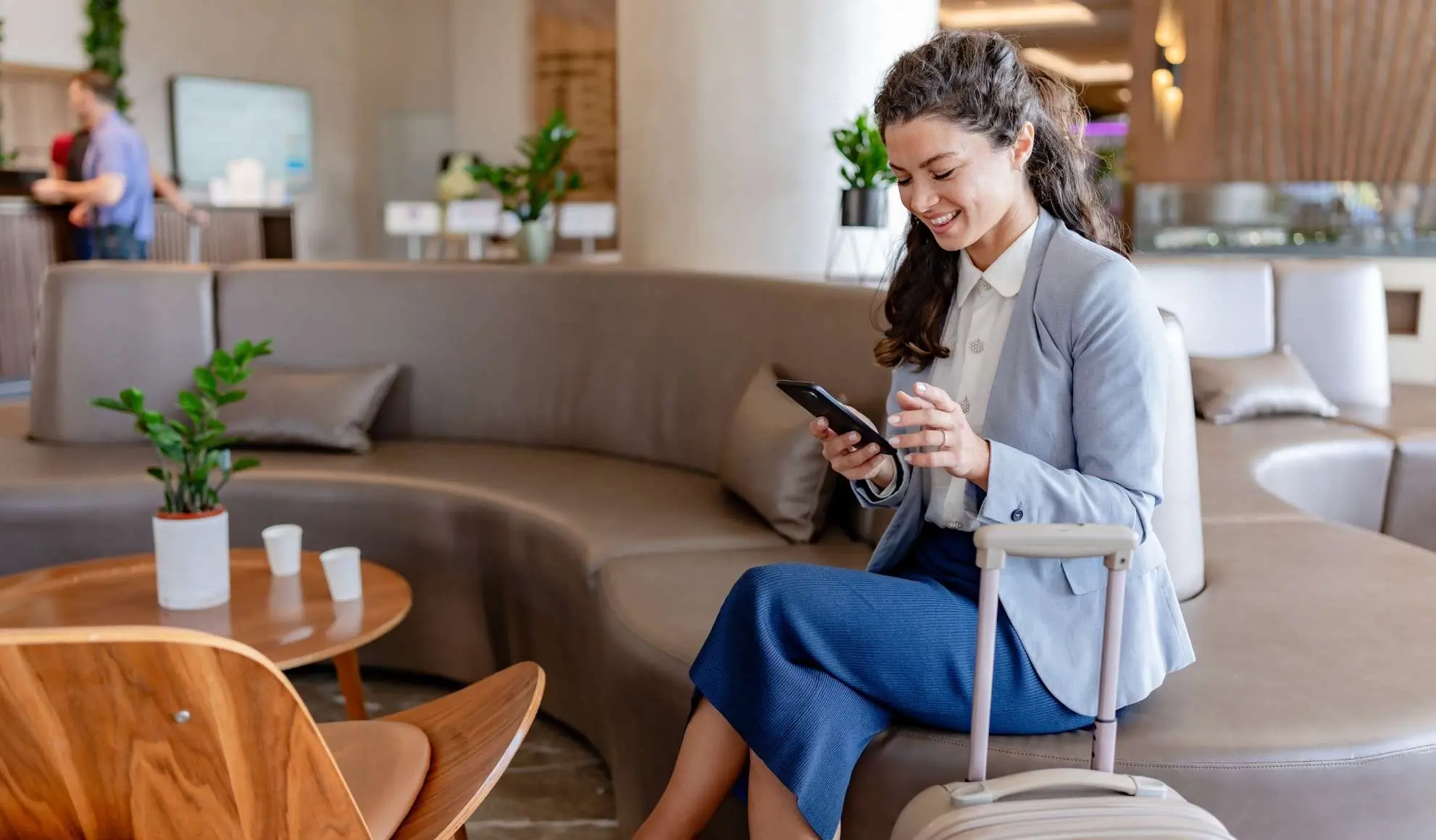 viaggio-bleisure-manager-lounge-hotel Professionista in lounge d’hotel con trolley e smartphone, esempio di viaggio bleisure tra lavoro e relax. Flyness, air chartering