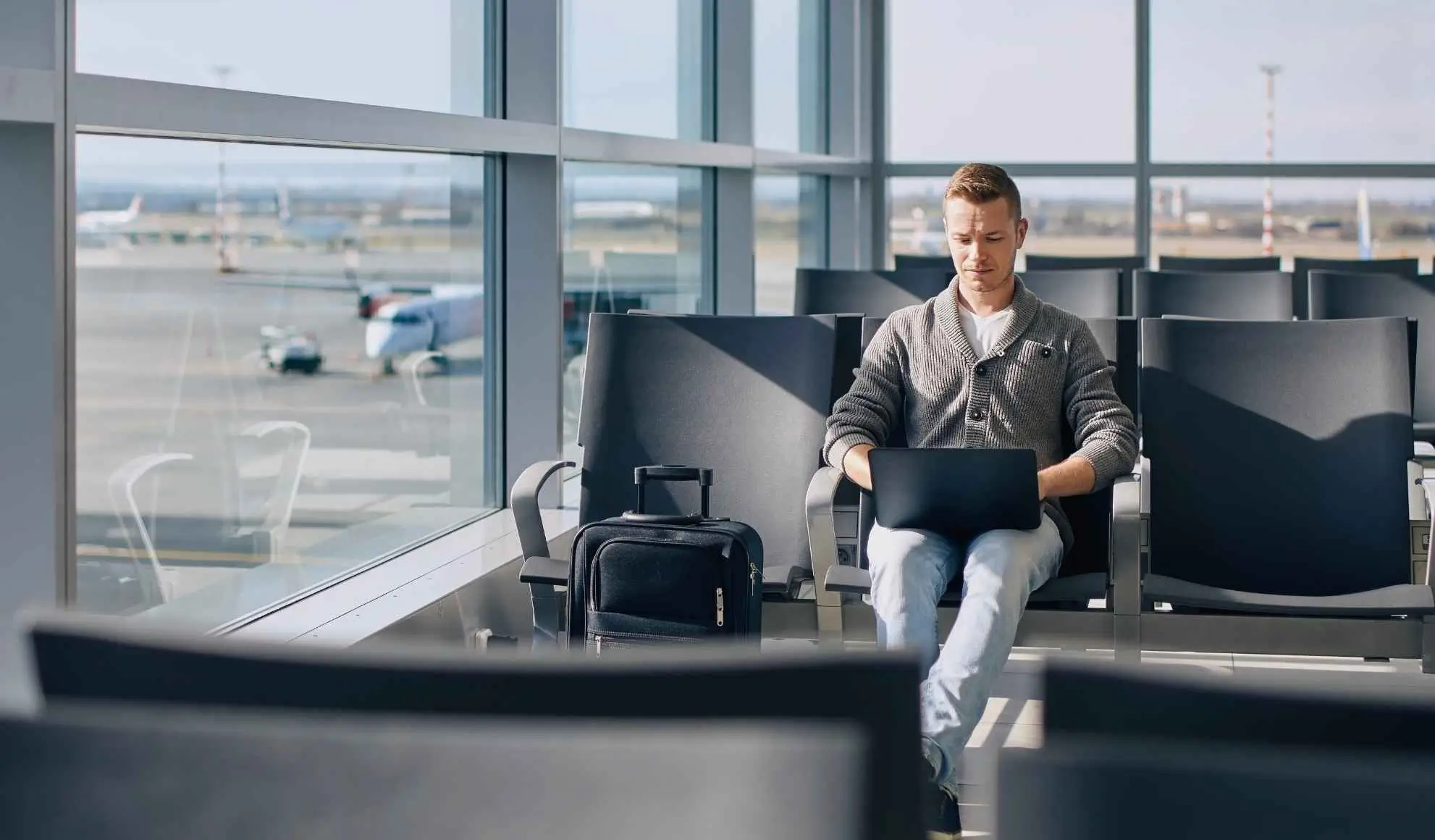 Travel Manager seduto nell’area d’attesa di un aeroporto mentre lavora al laptop, con un bagaglio accanto e aerei visibili dalla finestra.
